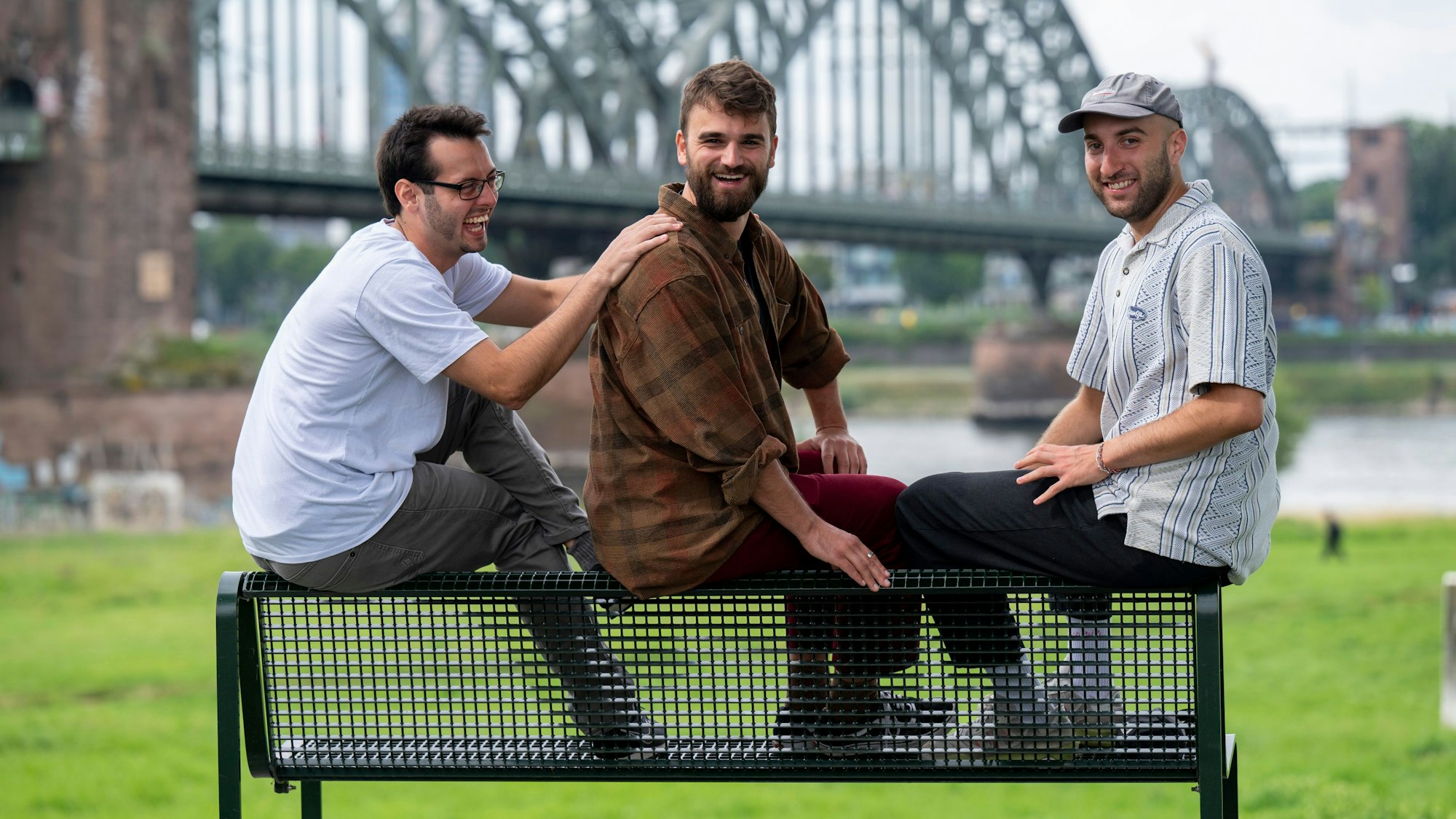 Leonard Träger, Simon Heuser und Andreas Stickelmann sitzen auf einer Bank am Rheinufer.