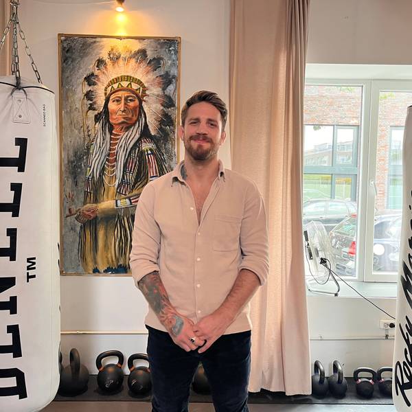 Ein Mann mit Bart steht vor einem Bild von einem Indianerhäuptling.