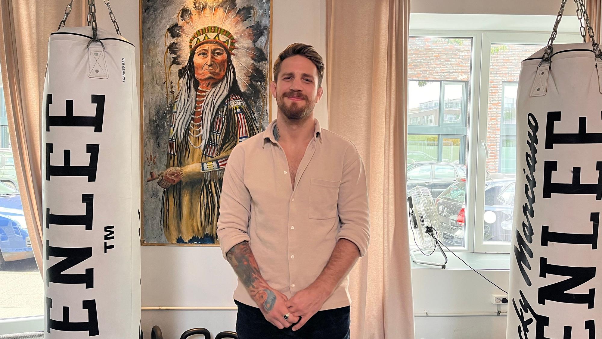 Ein Mann mit Bart steht vor einem Bild von einem Indianerhäuptling.