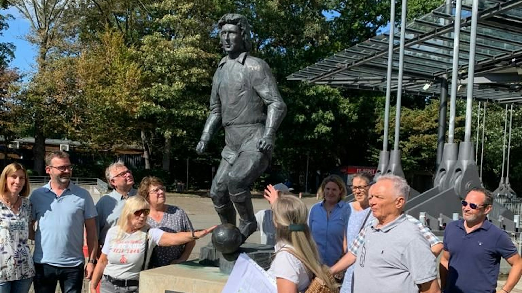 Vor dem Denkmal von Heinz Flohe referiert Cornelia Breuer (vorne) über die Geschichte der Stadionbauten.