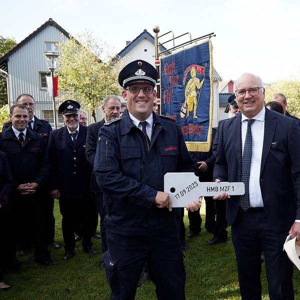 Bürgermeister Jochen Weiler und Löschgruppenführer Bernd Freuen halten den überdimensionalen Fahrzeugschlüssel. Hinter ihnen Feuerwehrleute mit einer Fahne und das zur Übergabe festlich geschmückte neue Fahrzeug.