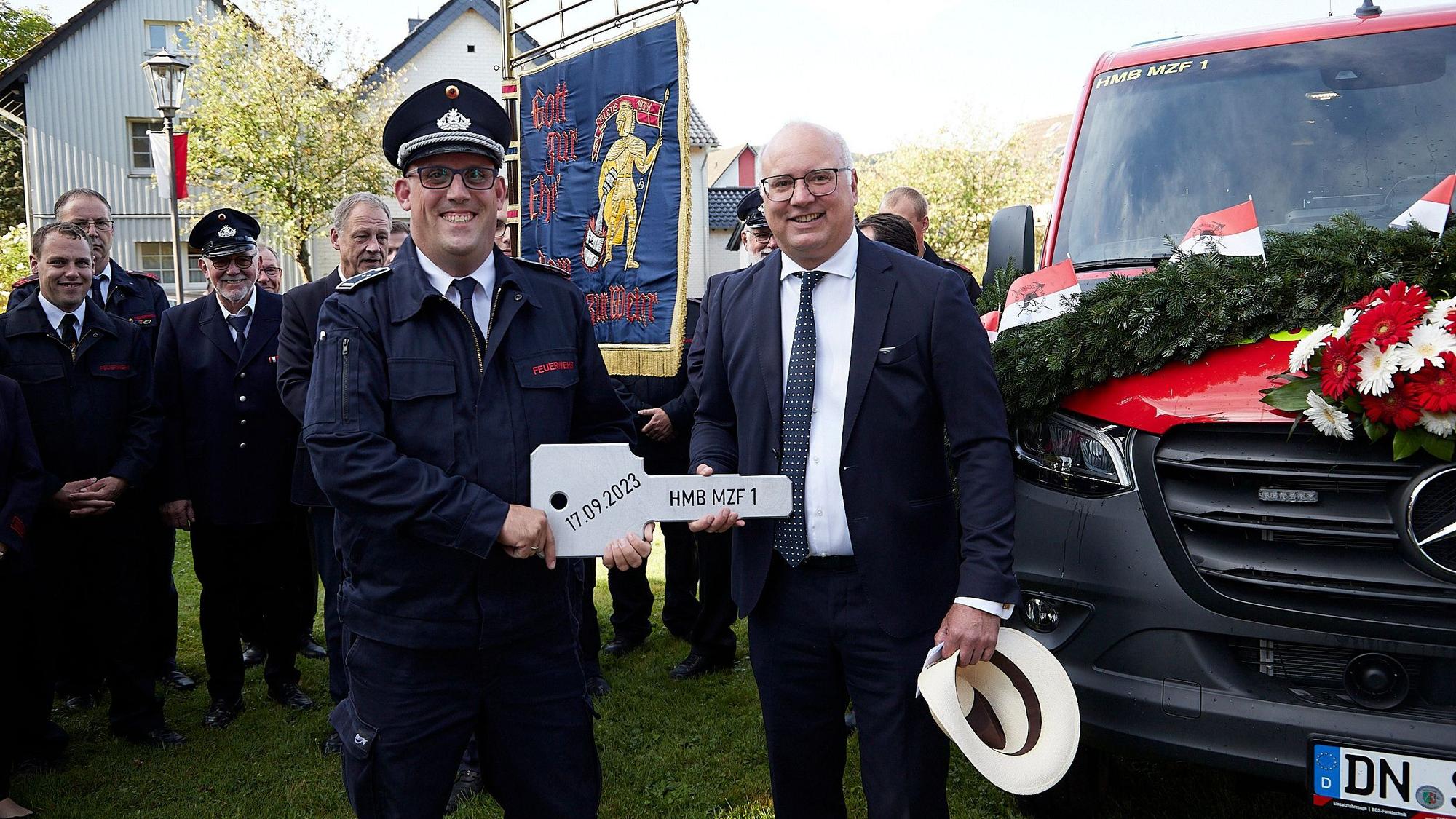 Bürgermeister Jochen Weiler und Löschgruppenführer Bernd Freuen halten den überdimensionalen Fahrzeugschlüssel. Hinter ihnen Feuerwehrleute mit einer Fahne und das zur Übergabe festlich geschmückte neue Fahrzeug.