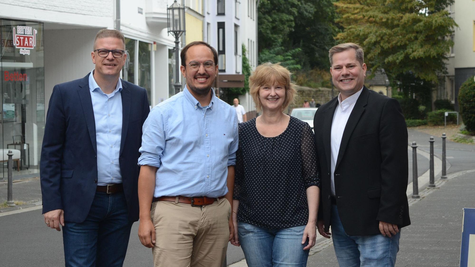 Drei Männer und eine Frau stehen auf dem Gehweg an einer Straße.