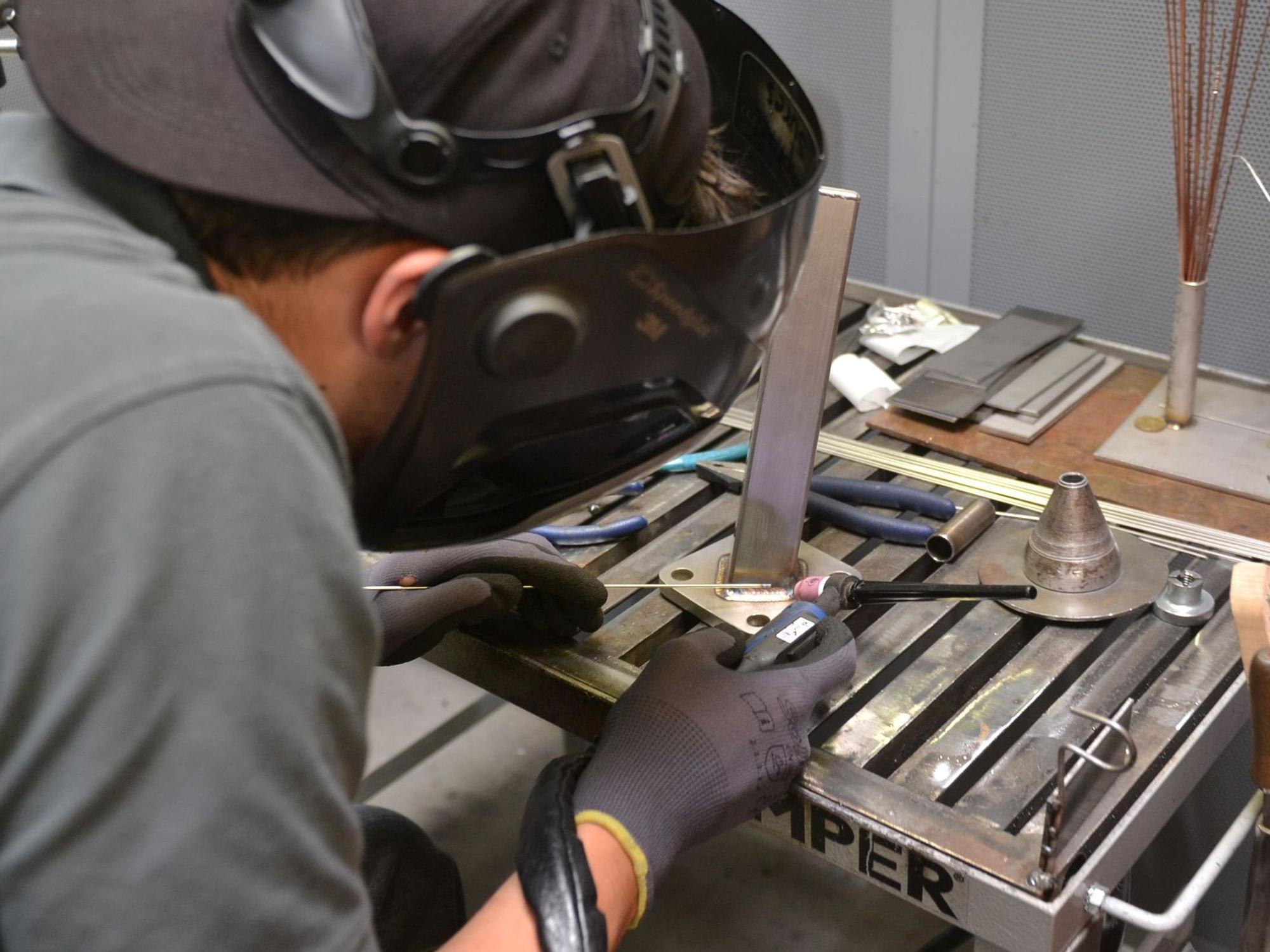 Ein junger Geselle ist von hinten zu sehen. Er trägt eine Schutzbrille und demonstriert, worauf es in seinem Beruf im Metallbau ankommt.