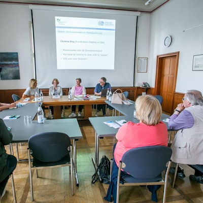 Mehrere Leute sitzen an Tischen und schauen auf eine Präsentation auf einer Leinwand.