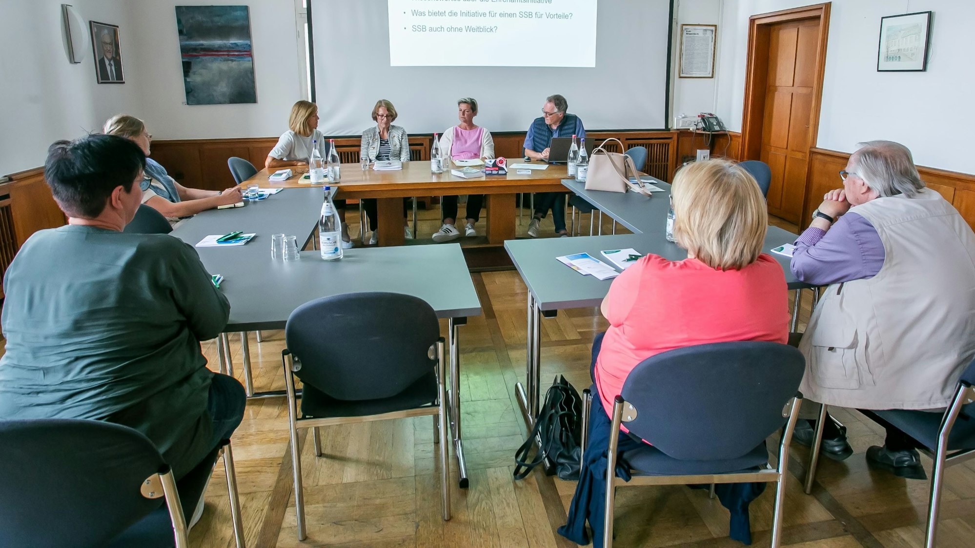 Mehrere Leute sitzen an Tischen und schauen auf eine Präsentation auf einer Leinwand.