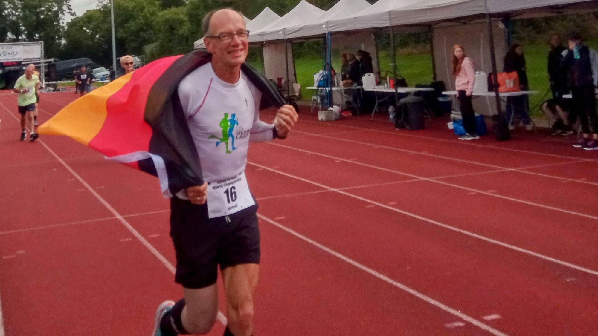 Ein Mann mit Halbglatze läuft auf einer roten Leichtathletikbahn. Er trägt eine Starternummer am Trikot hat und eine Deutschlandfahne um die Schultern.