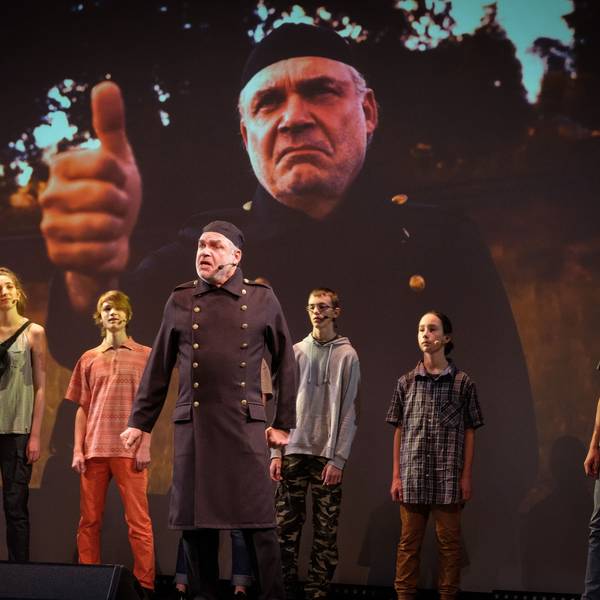 Darsteller eines Theaterstücks stehen vor einer Leinwand, auf der der Schauspieler in der Mitte noch einmal groß zu sehen ist.