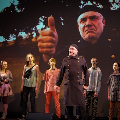 Darsteller eines Theaterstücks stehen vor einer Leinwand, auf der der Schauspieler in der Mitte noch einmal groß zu sehen ist.