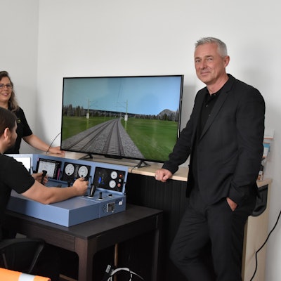 Vier Personen stehen am Simulator eines Lok-Führerstandes. Einer von ihnen steuert die Lok über eine Strecke, die auf dem Bildschirm zu sehen ist.