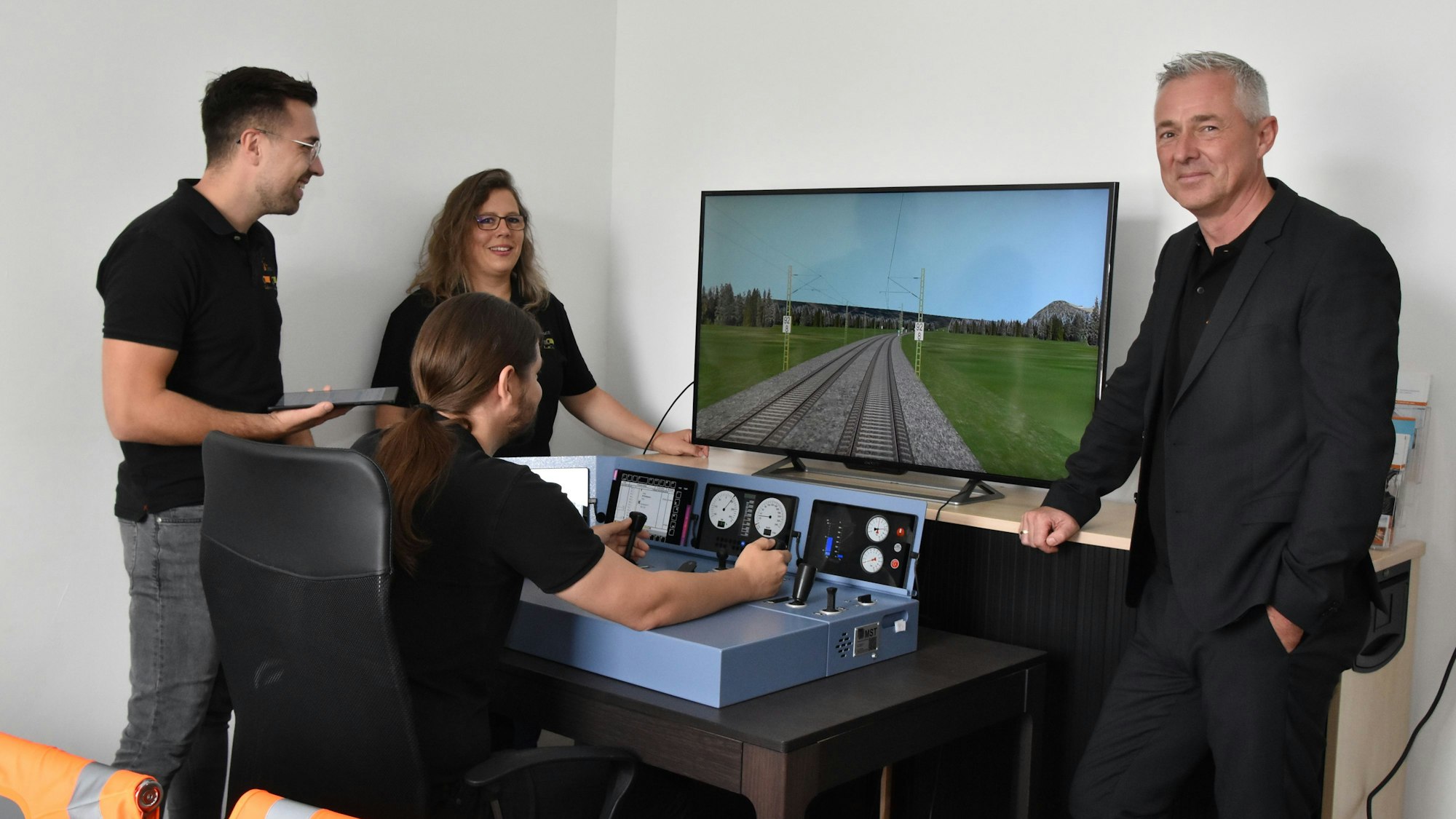 Vier Personen stehen am Simulator eines Lok-Führerstandes. Einer von ihnen steuert die Lok über eine Strecke, die auf dem Bildschirm zu sehen ist.