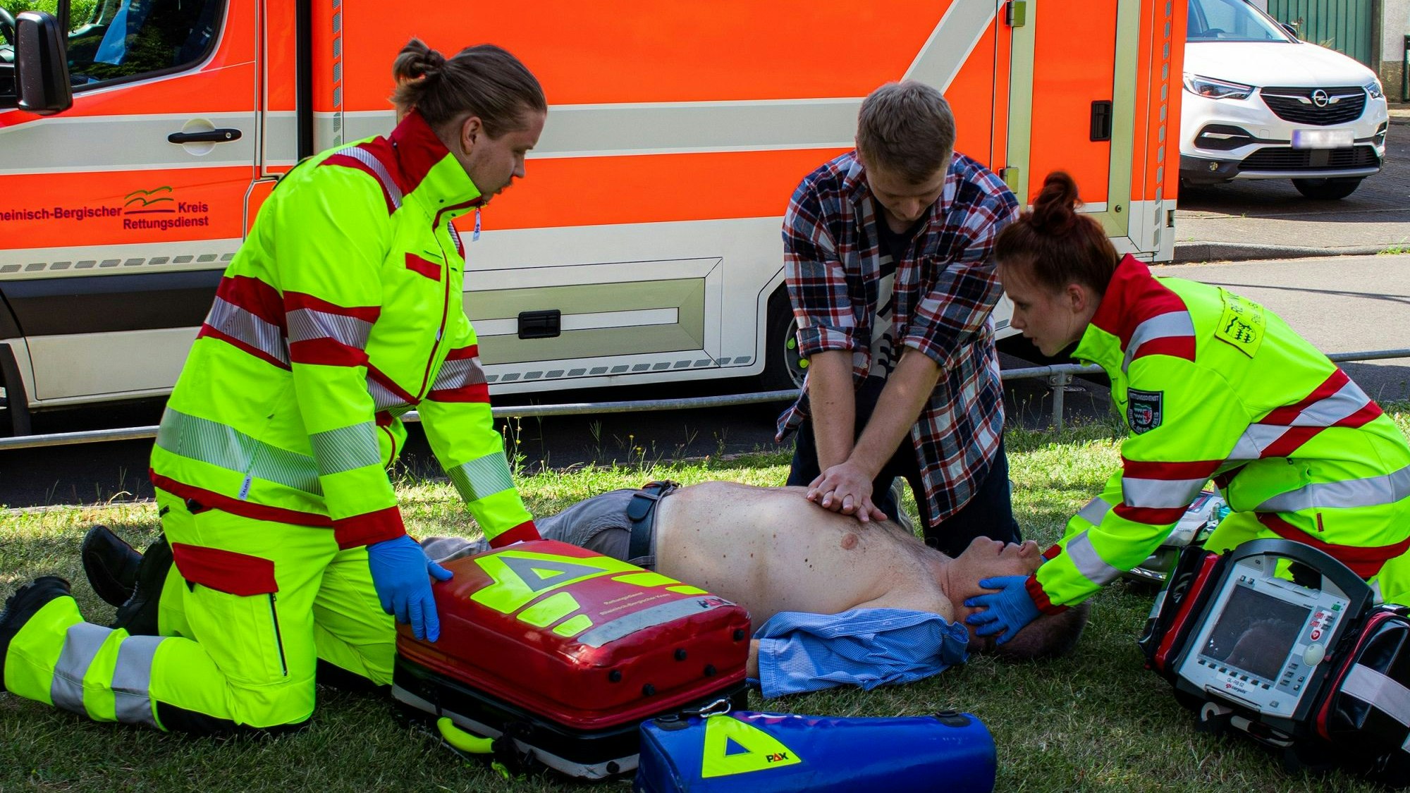 Ein Ersthelfer macht Herz-Druckmassage bei einem Patienten, zwei Rettungssanitäterinnen sind dabei,