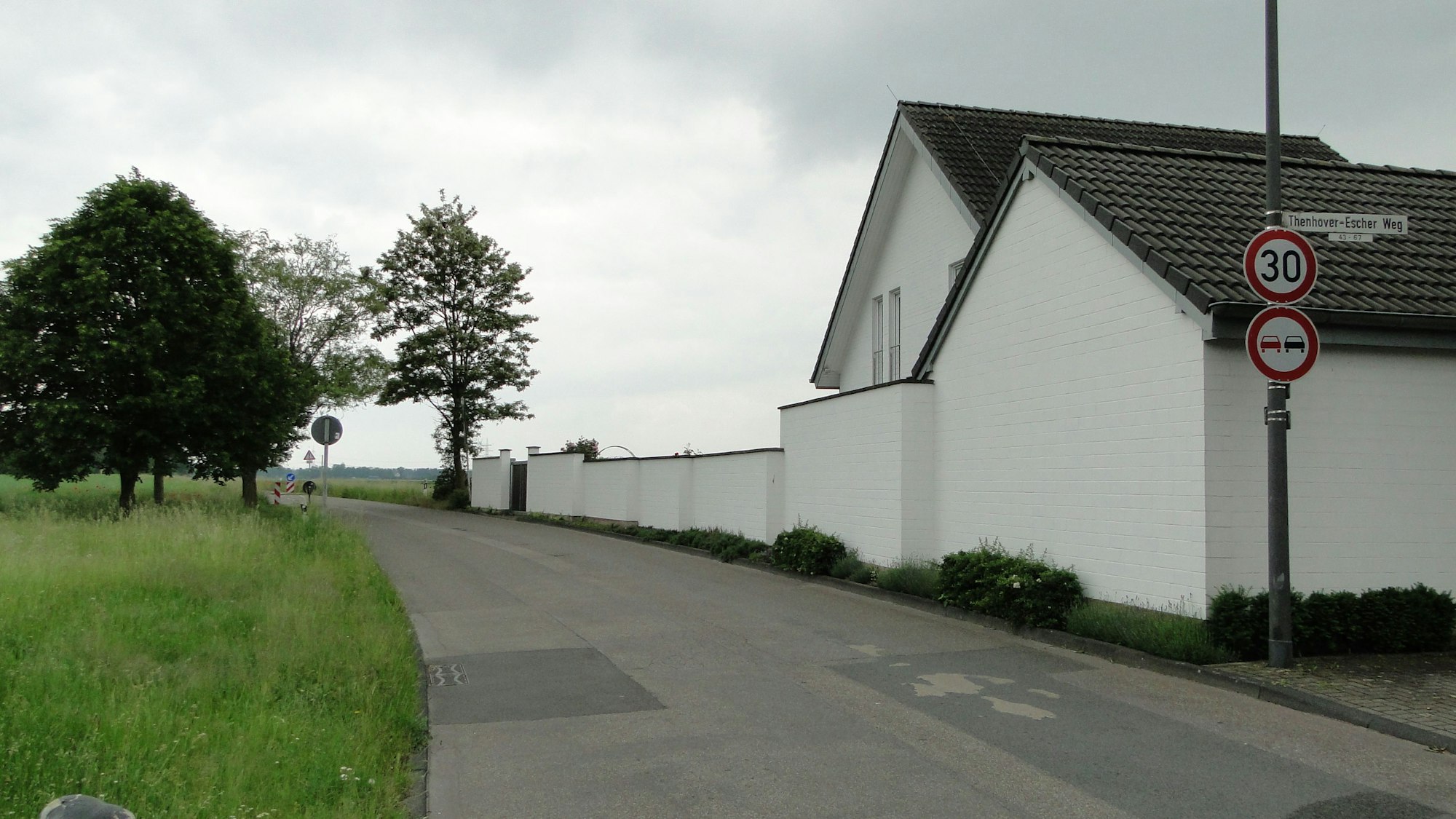 An einer Landstraße liegt ein weißes Haus mit Mauer und auf der anderen Seite eine Wiese mit vereinzeltem Baumbestand.