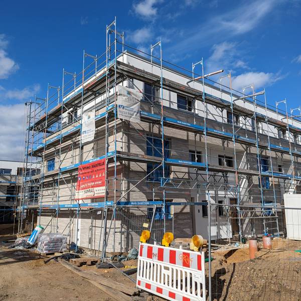 Ein Haus in der Bauphase. Rund um das Haus ist ein Gerüst aufgebaut. Im Hintergrund weitere Häuser mit Gerüsten.