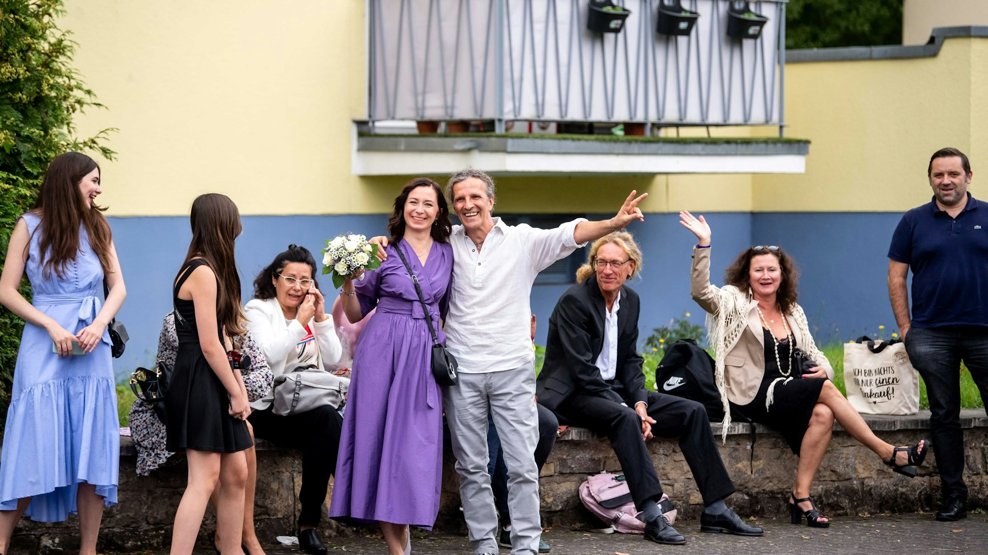 Das Bild zeigt das frisch getraute Ehepaar, das für die Kamera posiert. Ein Teil der Hochzeitsgäste sitzt im Hintergrund auf einer Mauer.