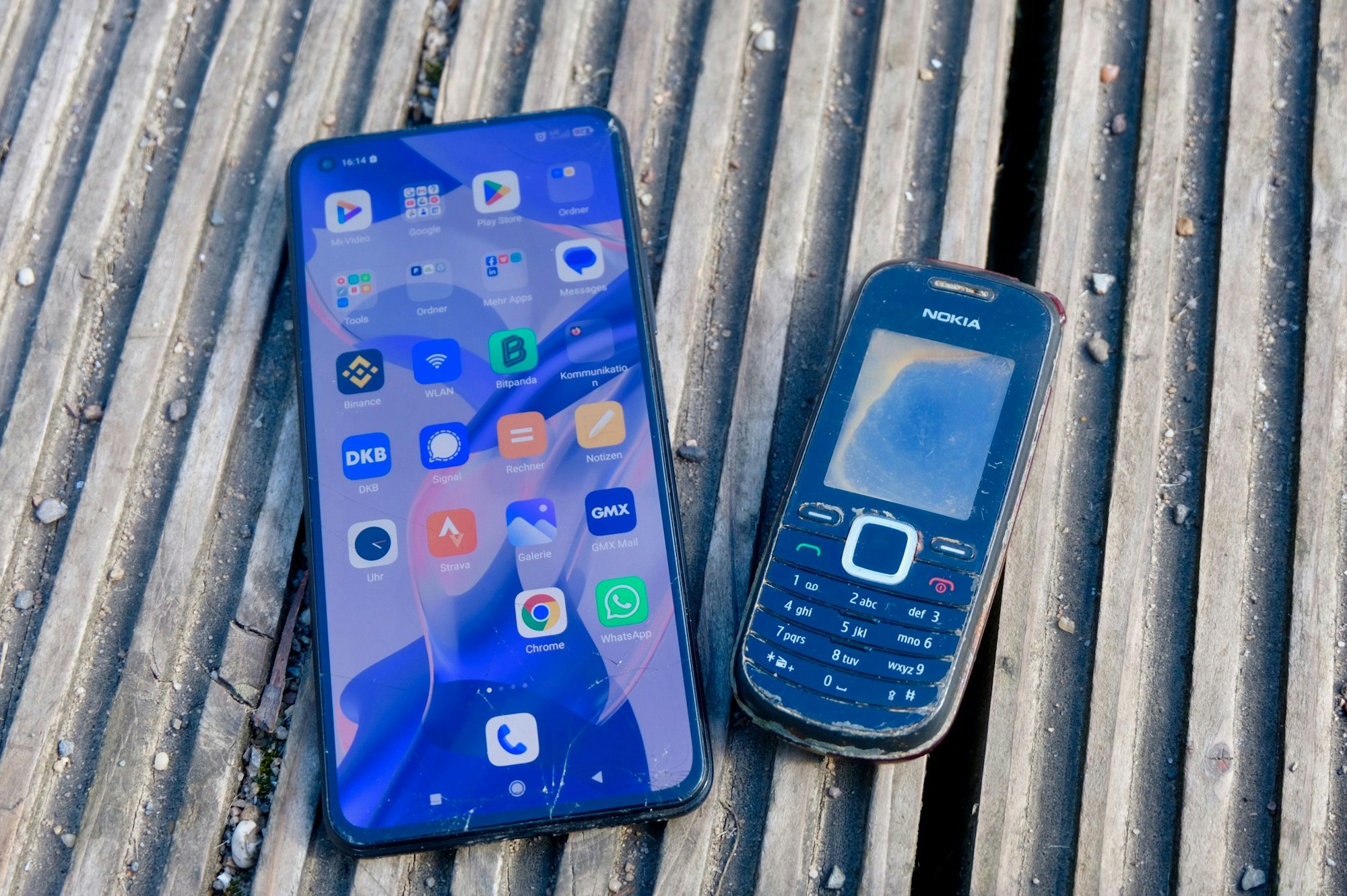Auf einem Holzsteg liegen zwei Mobiltelefone nebeneinander, ein modernes Smartphone und ein altes Nokia-Modell.
