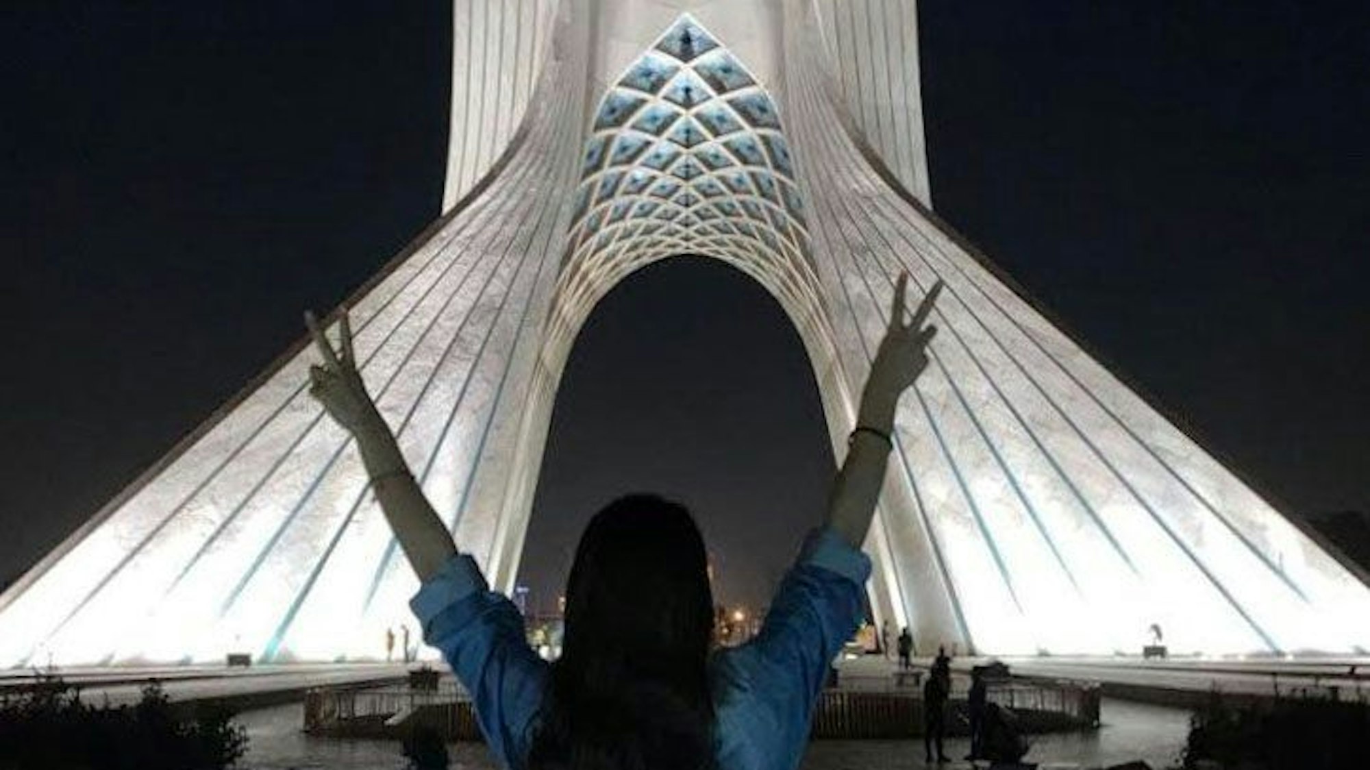 Eine junge Frau steht ohne das vorgeschriebene Kopftuch vor dem Azadi-Turm (Freiheitsturm) und zeigt mit beiden Händen das Sieges-Zeichen.
