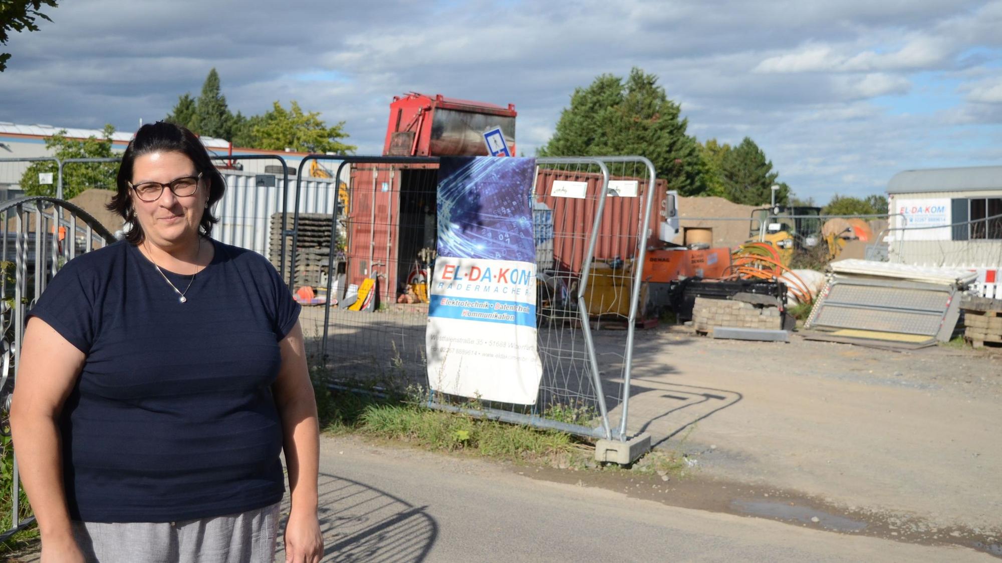 Eine Frau im blauen T-Shirt steht vor dem Grundstück, auf dem Baumaschinen geparkt werden. Im Hintergrund stehen Container und ein Bauwagen.