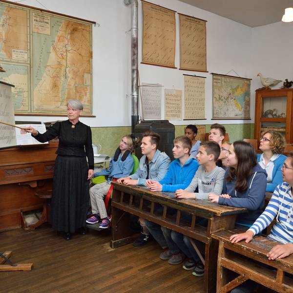 Jugendliche sitzen auf historischen Schulbänken aus Holz. An der Tafel steht eine als Lehrerin in der früheren Zeit in einem schwarzen langen Kleid verkleidete Mitarbeiterin des Museums.
