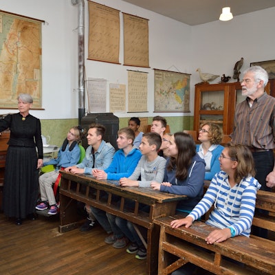 Jugendliche sitzen auf historischen Schulbänken aus Holz. An der Tafel steht eine als Lehrerin in der früheren Zeit in einem schwarzen langen Kleid verkleidete Mitarbeiterin des Museums.