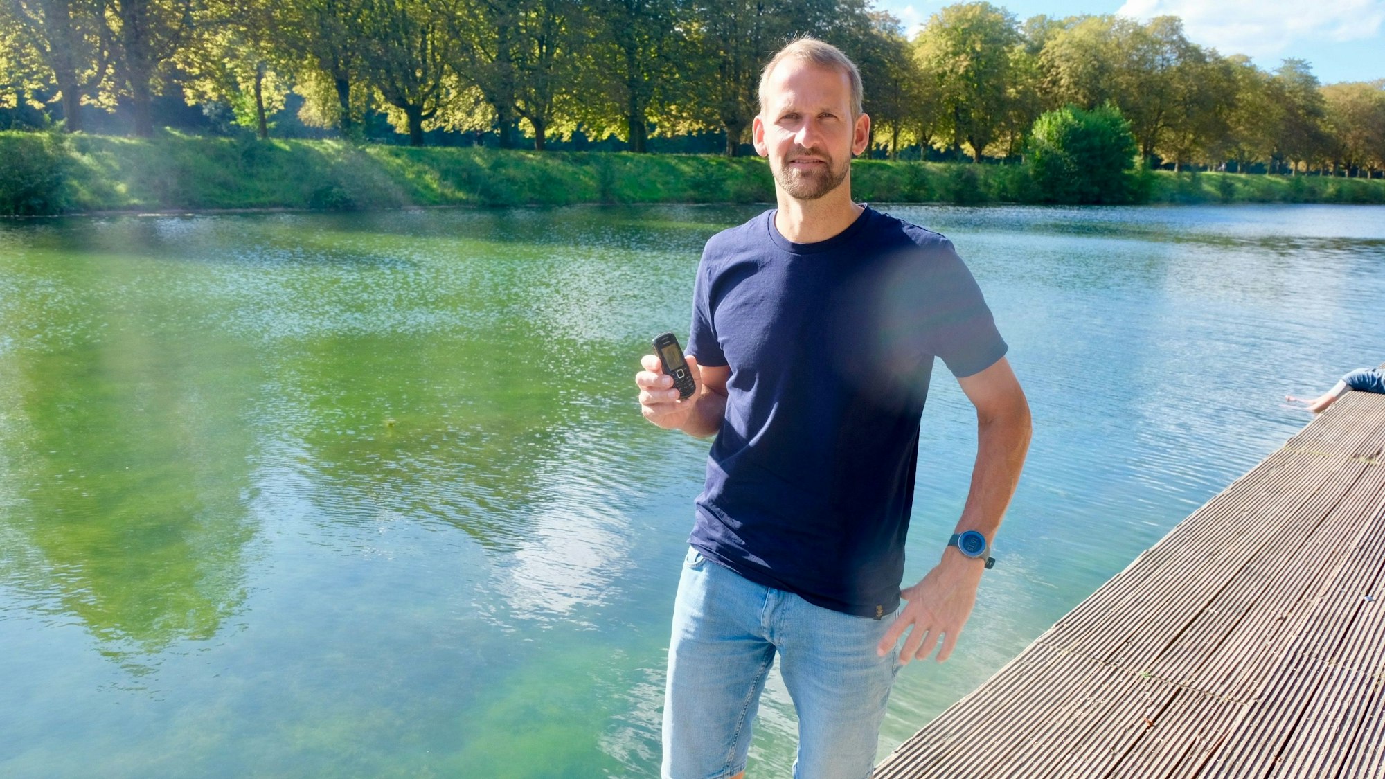 Ein Mann steht auf einem Steg an einem See, in der Hand hält er ein kleines Telefon.