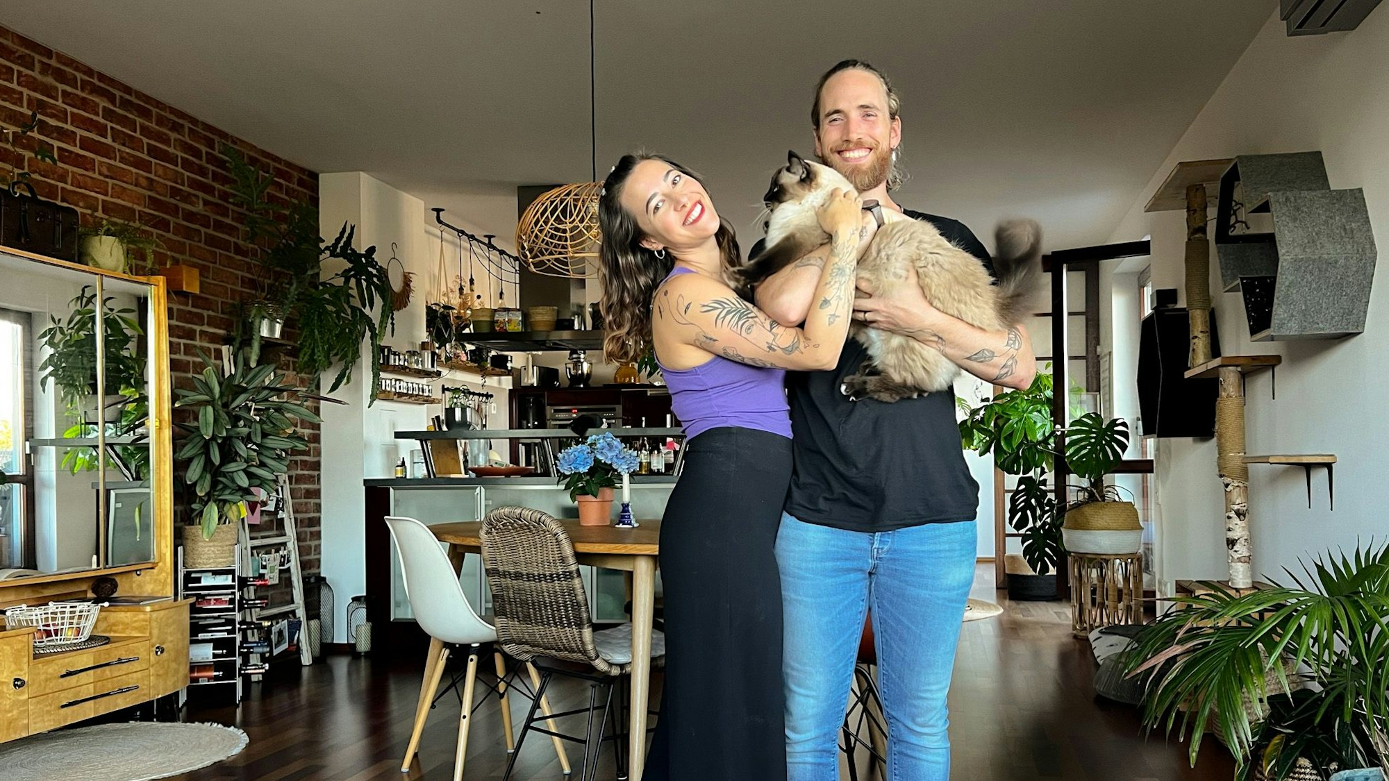 Eine Frau und ein Mann stehen in einem Wohnraum und lachen in die Kamera. Er hat eine Katze auf dem Arm.