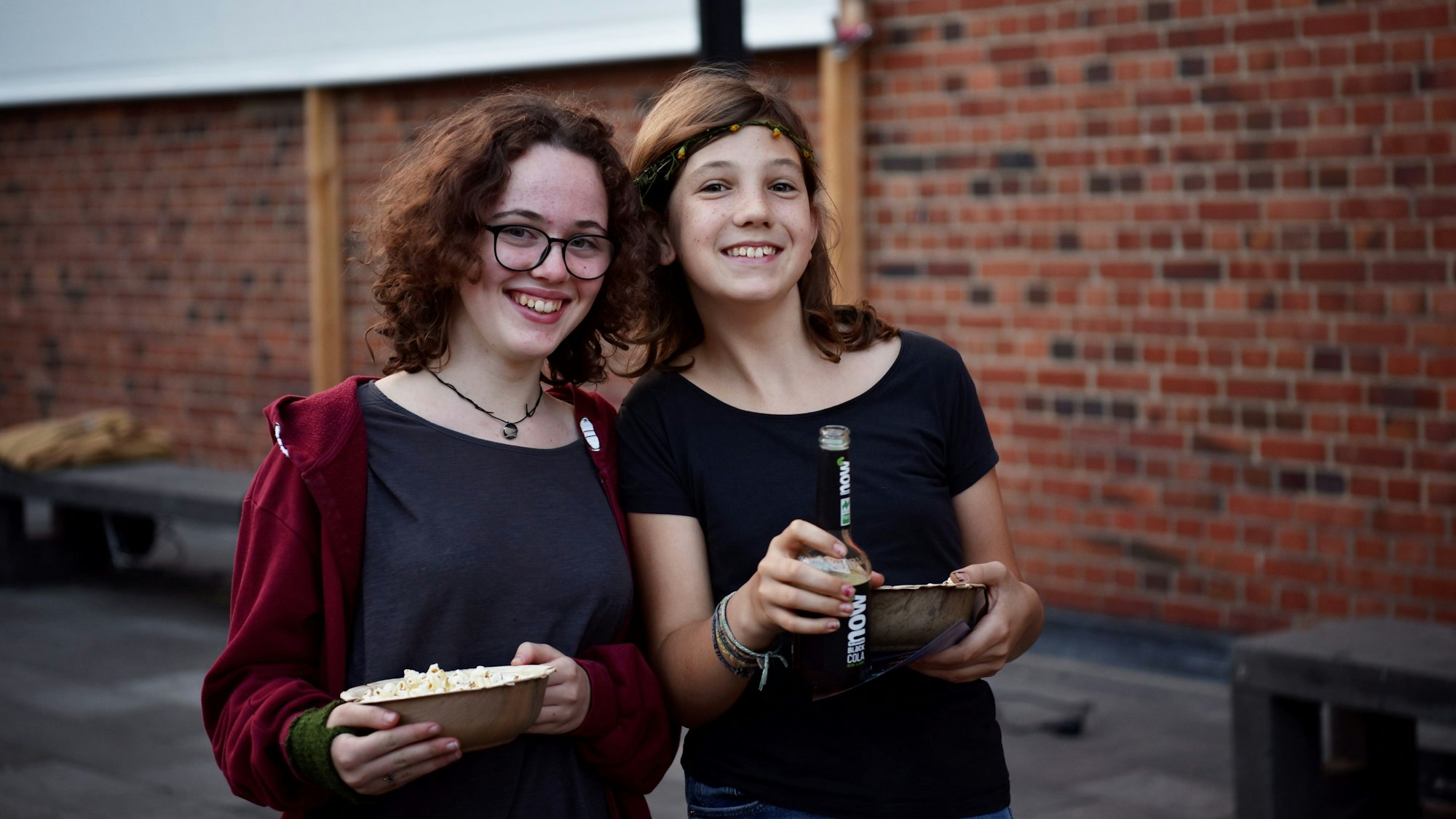 Die beiden Mädchen stehen nebeneinander. Sie haben Schalen mit Popcorn und eine Flasche Cola in der Hand.