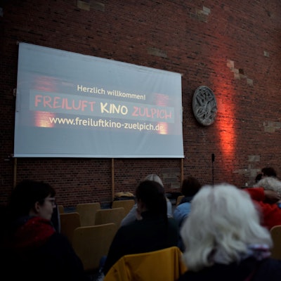 Das Foto zeigt die Leinwand auf dem Quiriniusplatz in Zülpich, davor Zuschauer. Das kostenlose Freiluftkino Zülpich findet immer im Frühjahr und im Herbst auf dem Quiriniusplatz statt.