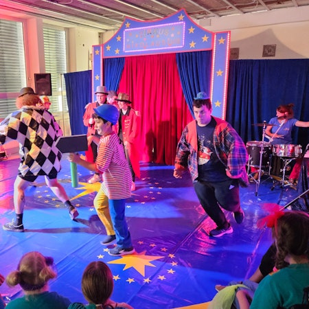 Bunt kostümierte Erwachsene und Jugendliche laufen in einer kleinen Zirkusmanege im Kreis.