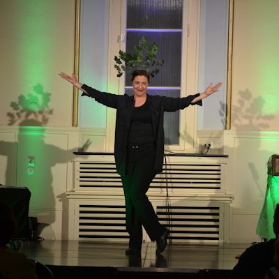 Die Kabarettistin steht in Schwarz gekleidet auf einer Bühne. Sie hat beide Arme ausgebreitet und lächelt.