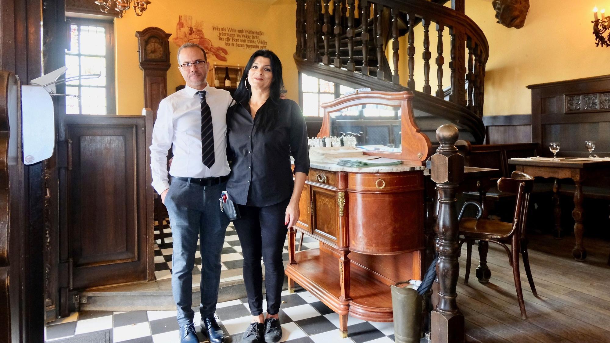 Ein Mann und eine Frau stehen nebeneinander in einem gediegenen Gastraum mit dunkler Holzvertäfelung und einem Fußboden im Schachbrett-Muster.