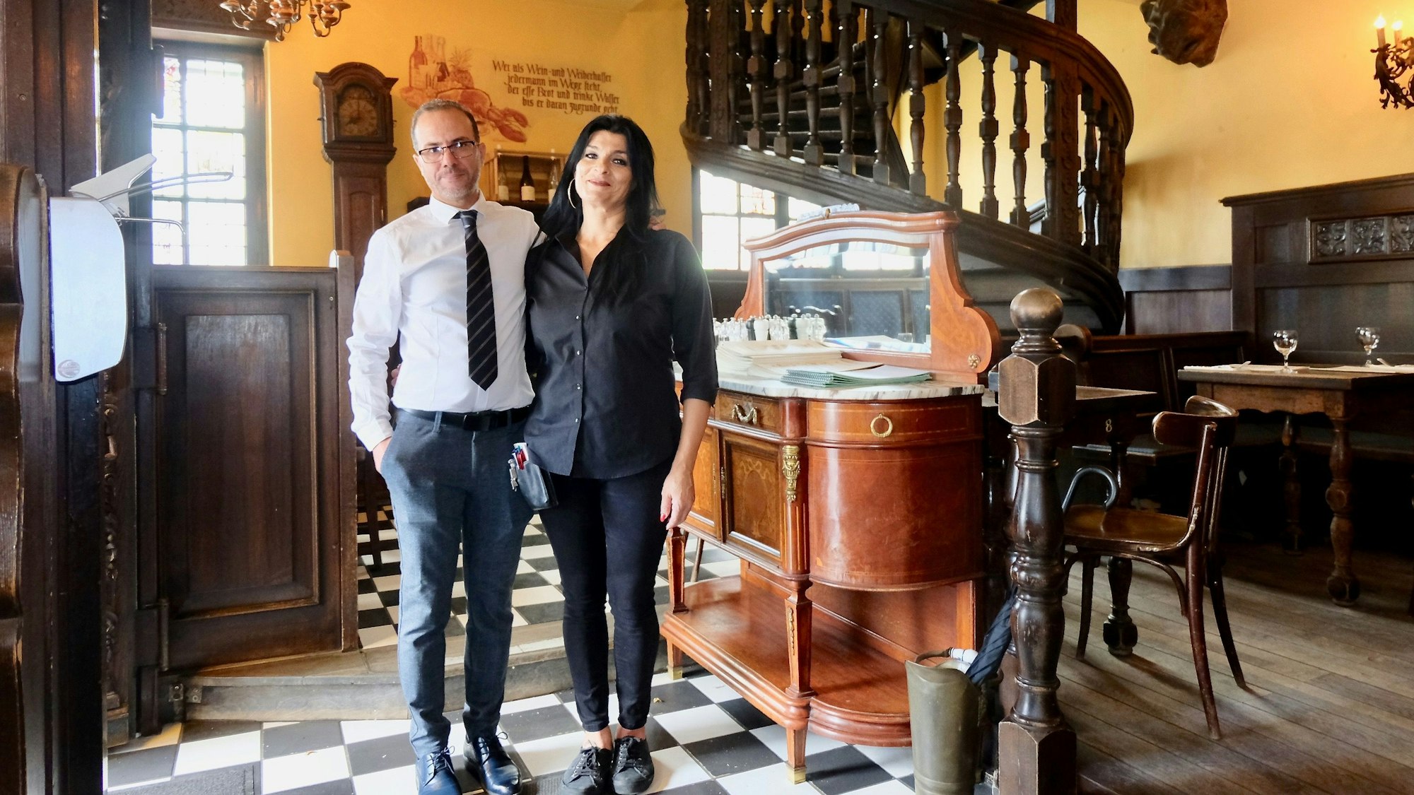 Ein Mann und eine Frau stehen nebeneinander in einem gediegenen Gastraum mit dunkler Holzvertäfelung und einem Fußboden im Schachbrett-Muster.