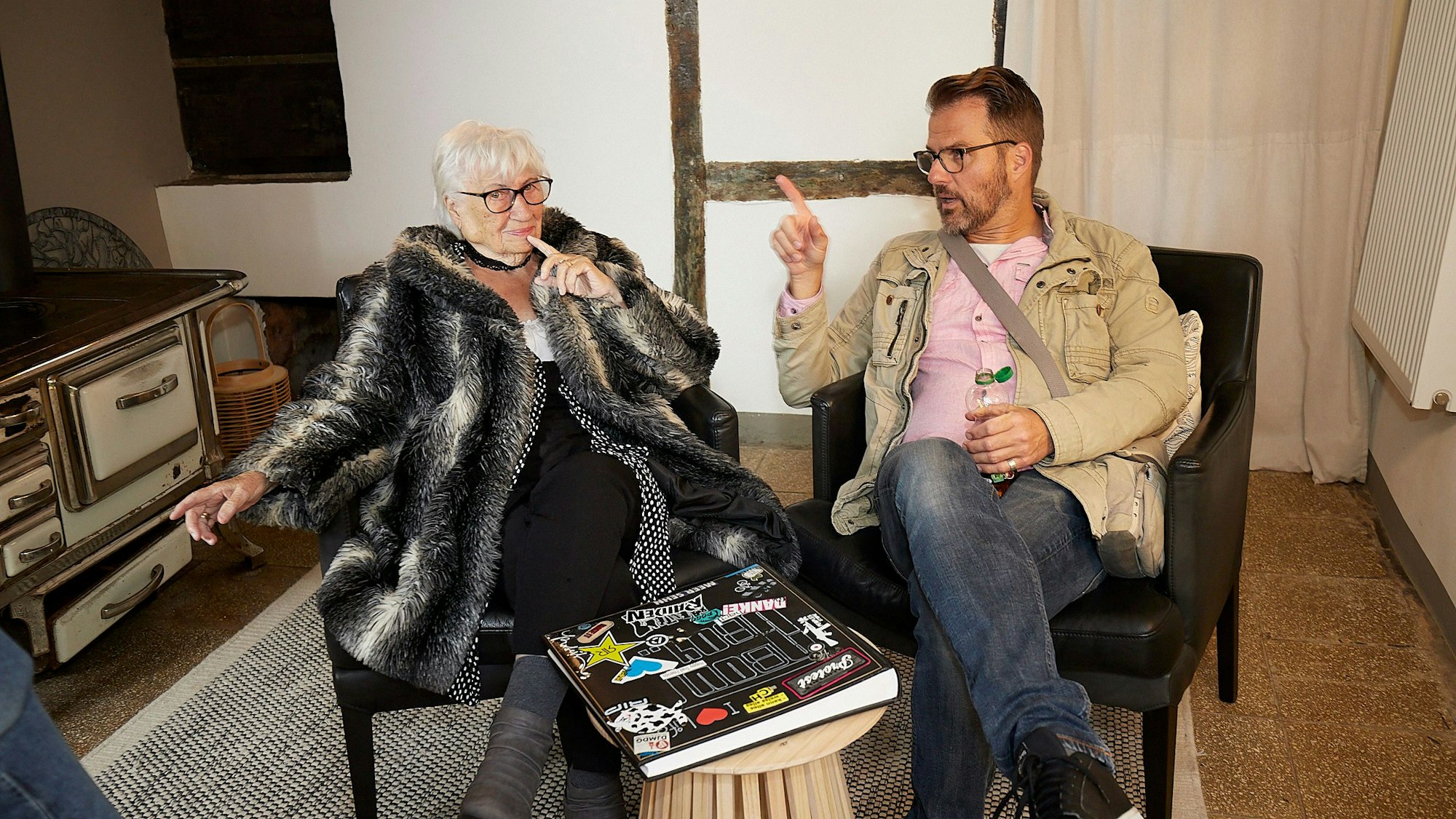 Ingo Wolfgarten und seine Großmutter Liesel Buchheldt sitzen in schwarzen Sesseln und unterhalten sich. Vor ihnen auf dem Tisch liegt ein Fotoalbum.