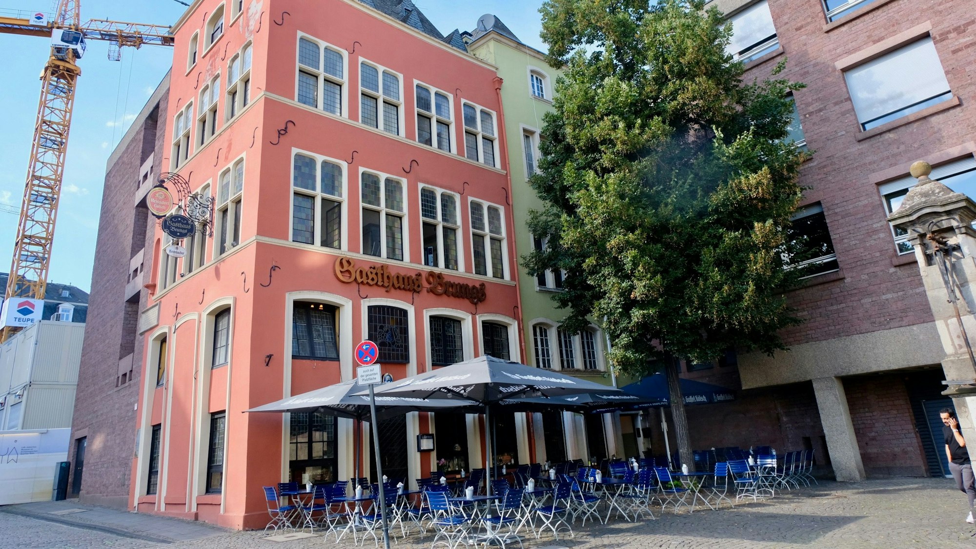 Hinter einer großen Terrasse mit Tischen, Stühlen und aufgespannten Sonnenschirmen stehen ein rotes und ein grünes Haus im klassischen Altstadt-Stil.