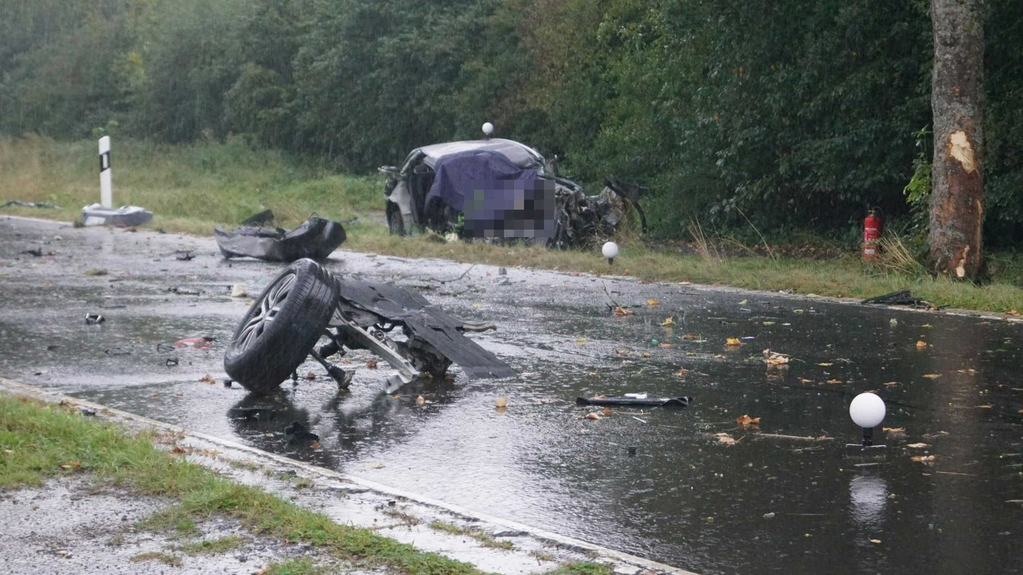 Das Foto zeigt die Unfallstelle mit dem zerstörten Wagen und dem Baum, auf den das Auto geprallt ist. Auf der Fahrbahn liegen abgerissene Fahrzeugteile.