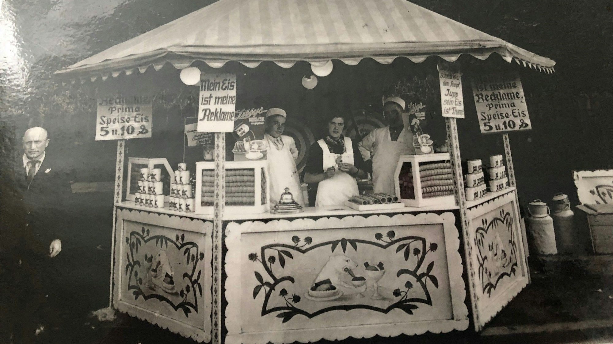 Auf dem Schwarz-Weiß-Foto stehen Frauen in weißen Schürzen in einem Pavillon, handgeschriebene Schilder preisen Eis an.