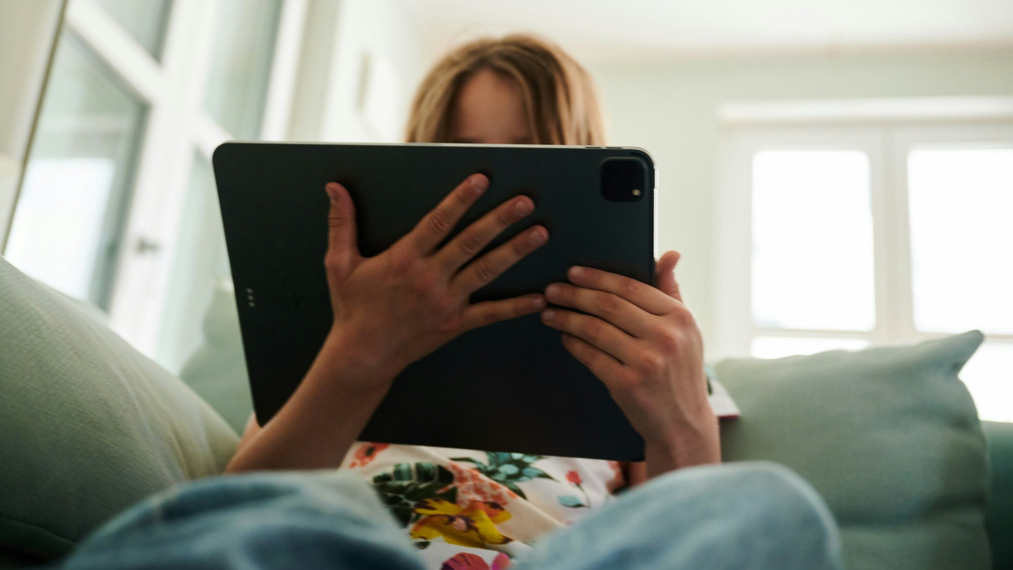 Ein Mädchen sitzt auf der Couch und guckt in das Tablet ihrer Eltern.