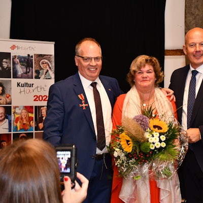 Ein Mann mit Orden am Revers, eine Frau mit Blumenstraße, und ein Mann um dunklen Anzug stehen auf einer Bühne.