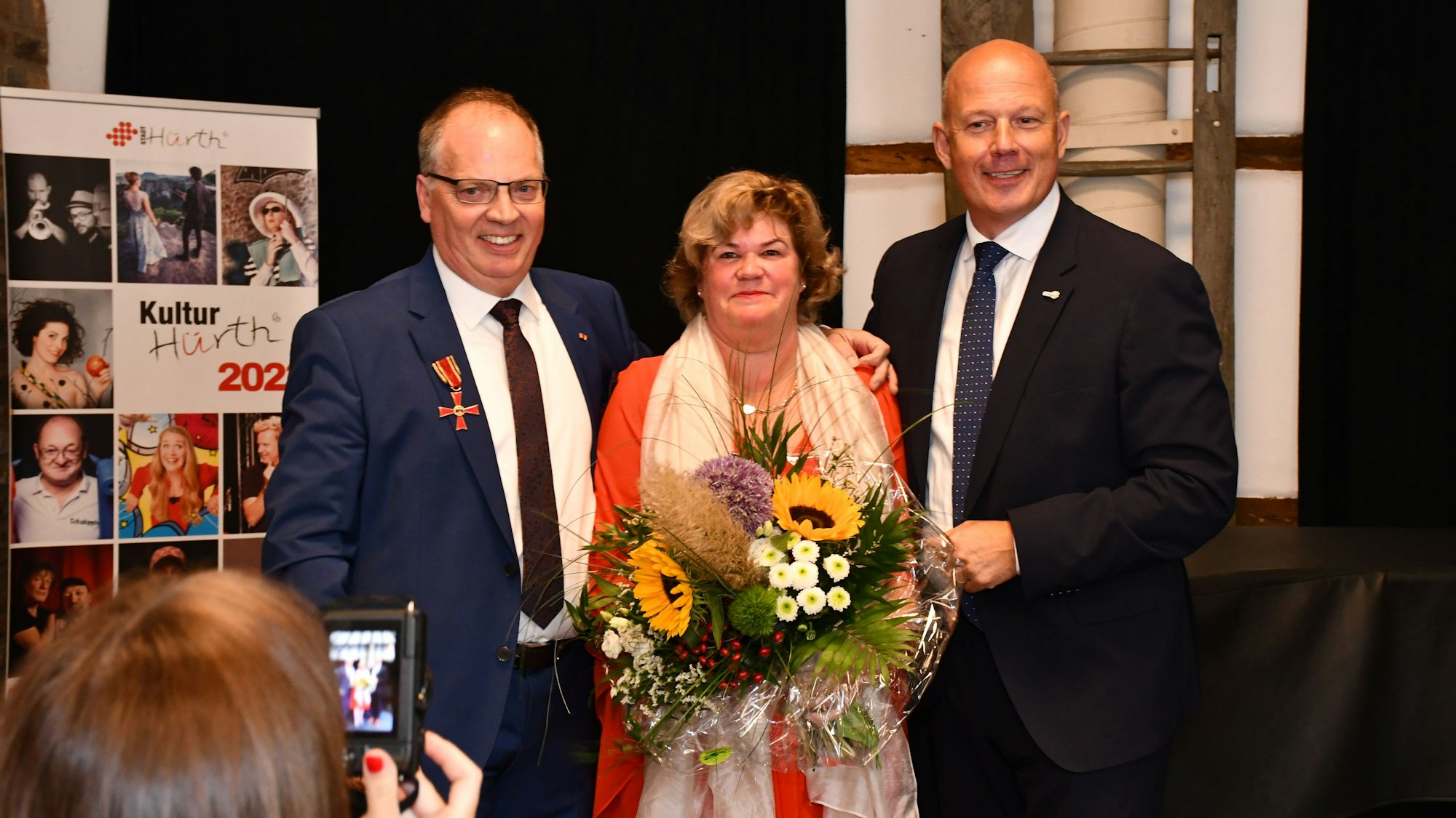 Ein Mann mit Orden am Revers, eine Frau mit Blumenstraße, und ein Mann um dunklen Anzug stehen auf einer Bühne.