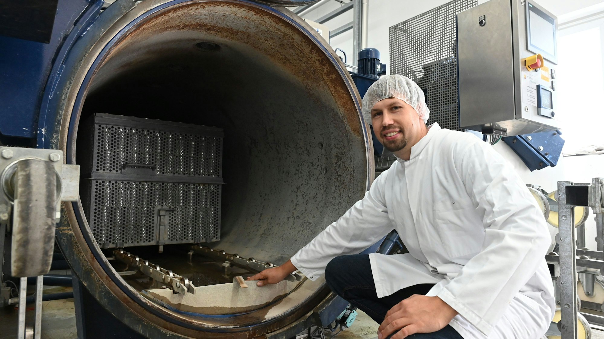 Markus Koller hockt vor einem geöffneten Autoklaven, in dem das Tierfutter gekocht wird. Im Inneren sind Dosen in einer Box zu sehen. Koller zeigt auf ein Schott, das er entwickelt hat, damit beim Öffnen so wenig Wasser wie eben möglich entweicht und es im Kreislaufprozess verbleiben kann.
