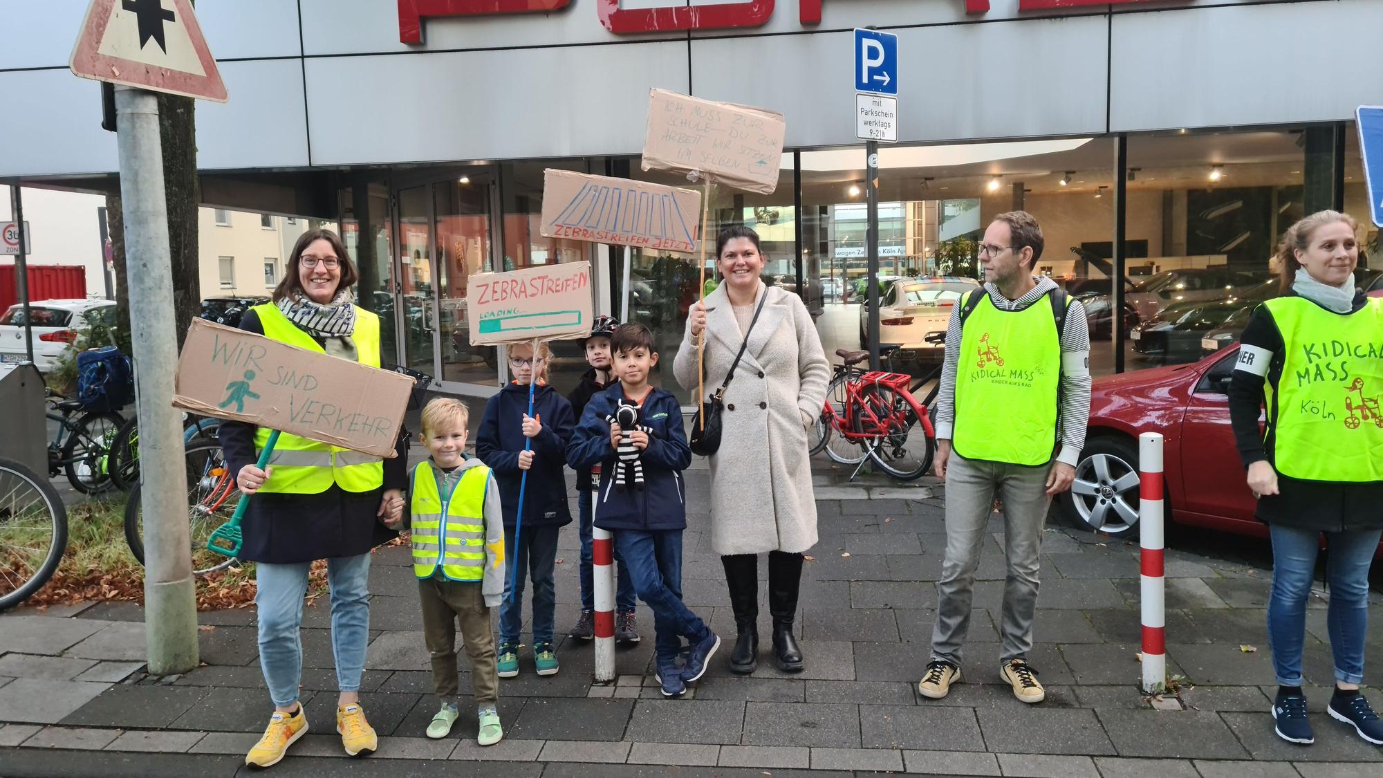Eltern und Kinder halten Pappschilder in die Höhe. Hinter ihnen ist das Autohaus Porsche zu sehen.