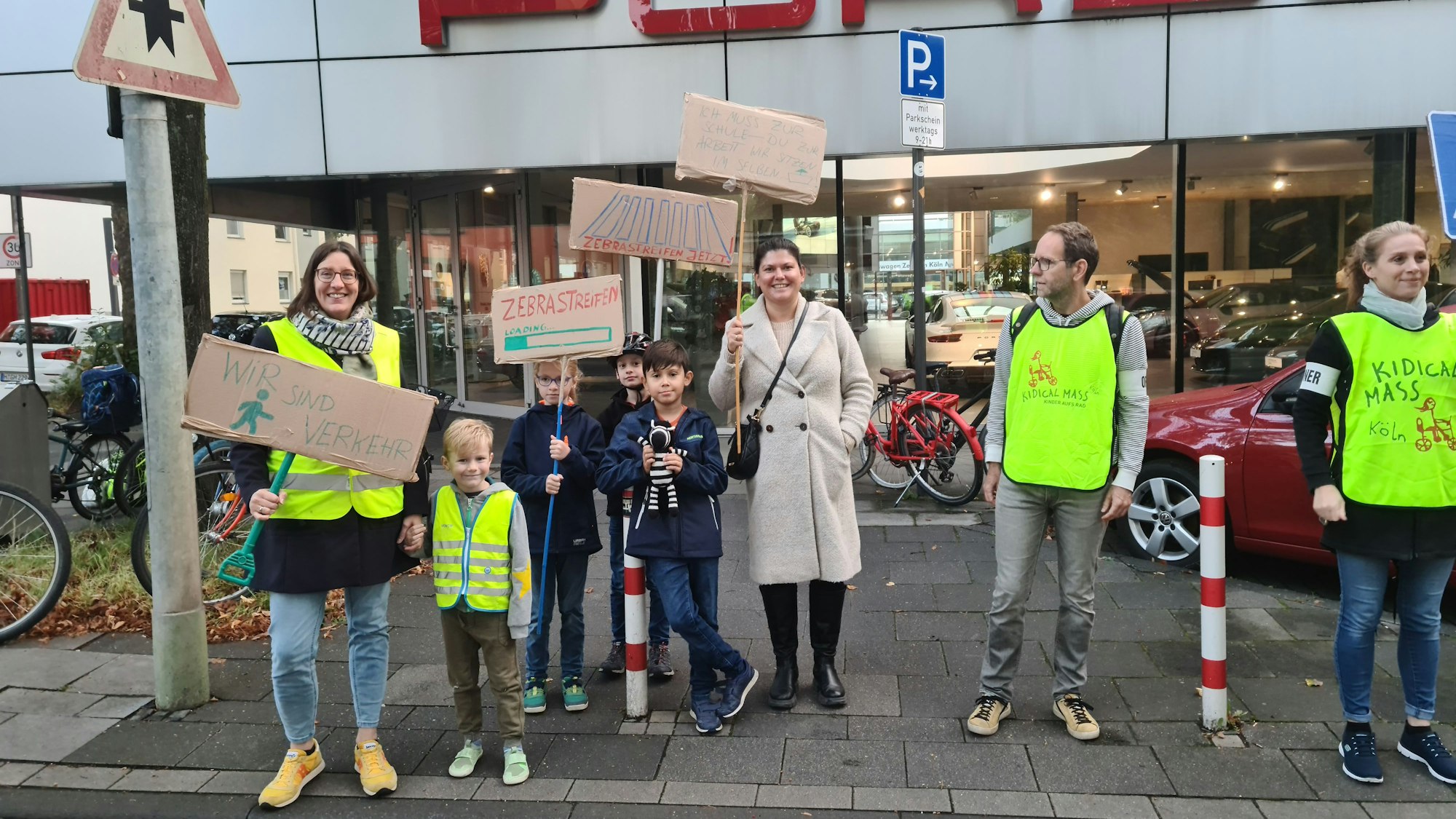 Eltern und Kinder halten Pappschilder in die Höhe. Hinter ihnen ist das Autohaus Porsche zu sehen.