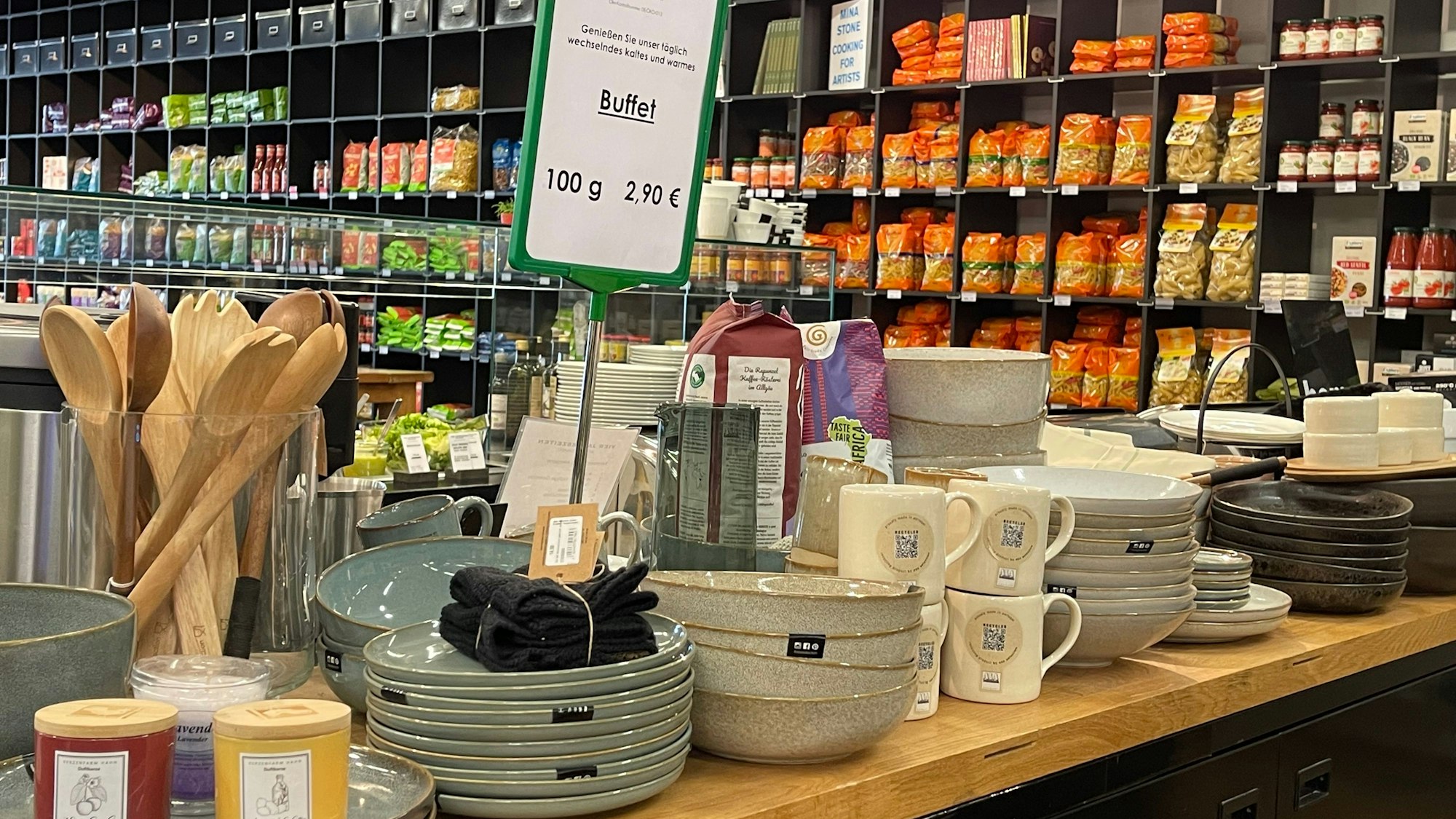 Ein Holztisch voll mit Tellern, Tassen und Löffeln, im Hintergrund steht ein langes, schwarzes Regal mit Nudelsorten.