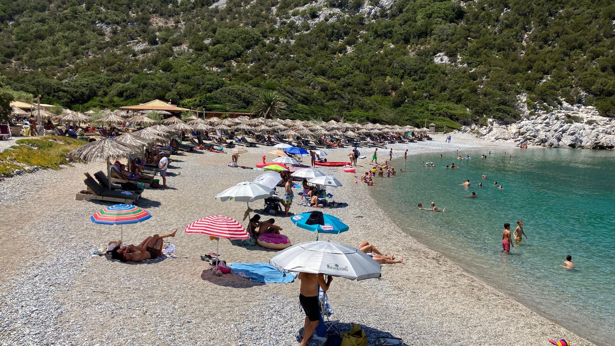 Sonnenschirme und Menschen am Strand, rechts ist das Meer zu sehen, im Hintergrund ein Berg