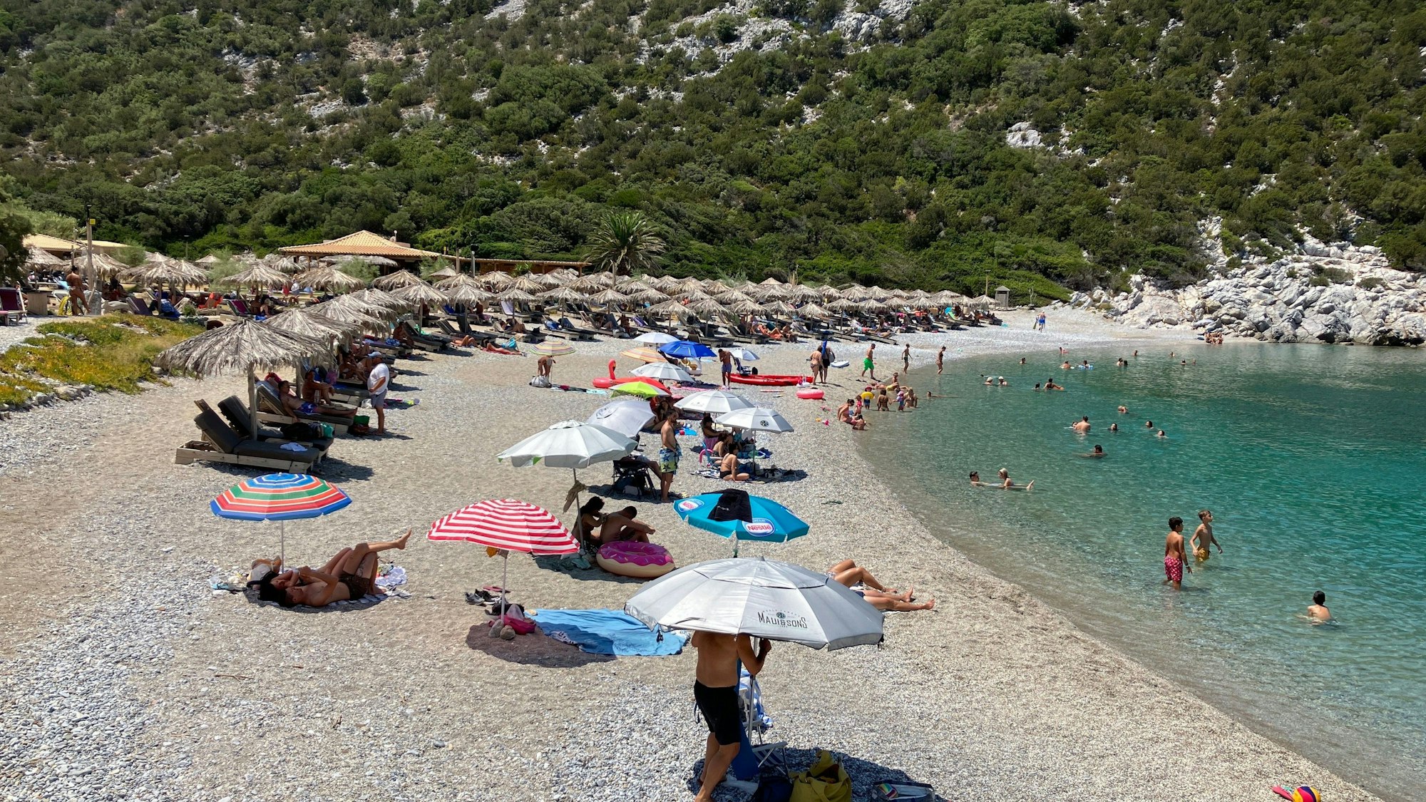 Sonnenschirme und Menschen am Strand, rechts ist das Meer zu sehen, im Hintergrund ein Berg