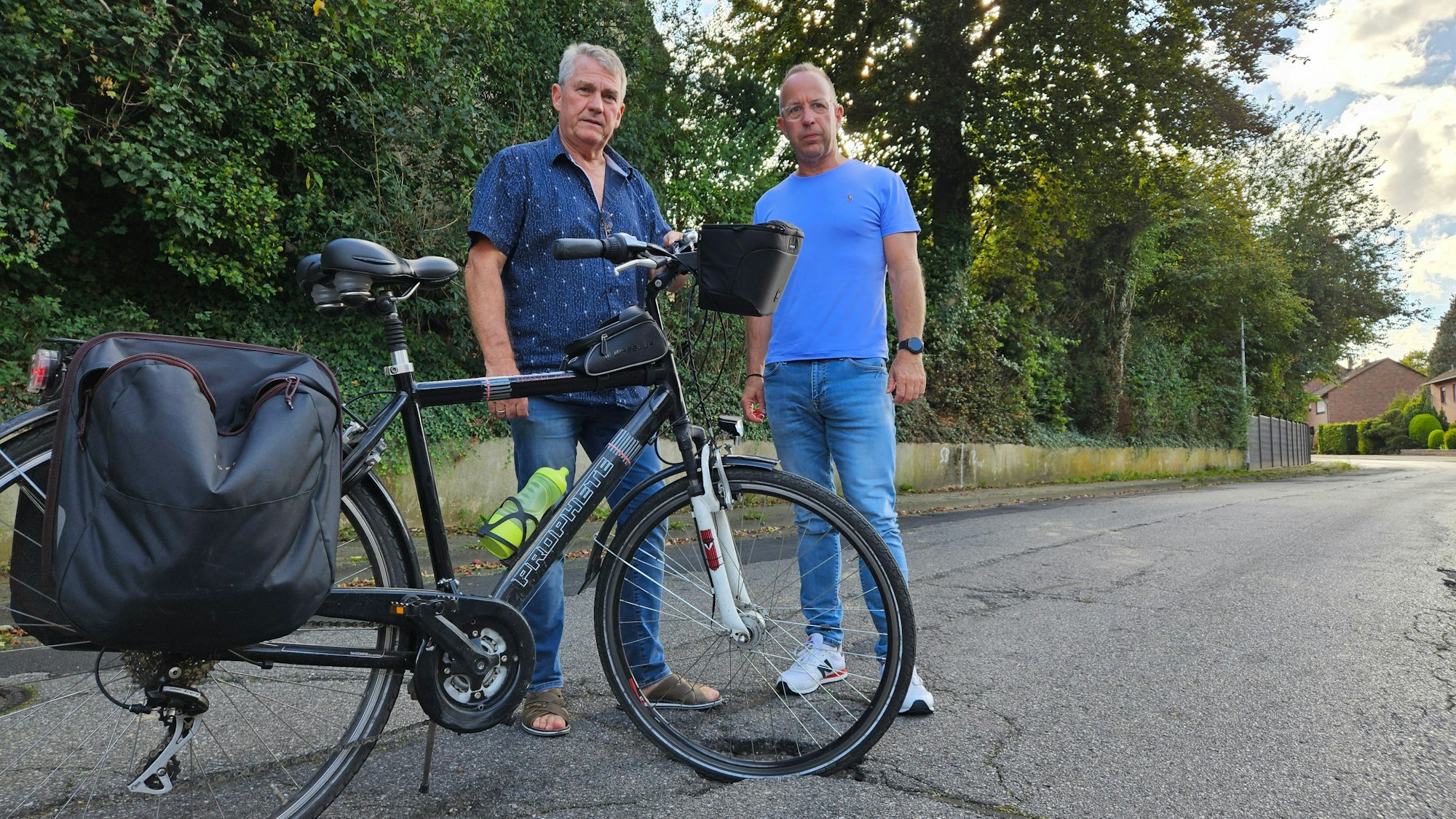 Zwei Männer stehen mit einem Fahrrad auf einer Straße, der Vorderreifen des Rads steht in einem Schlagloch.