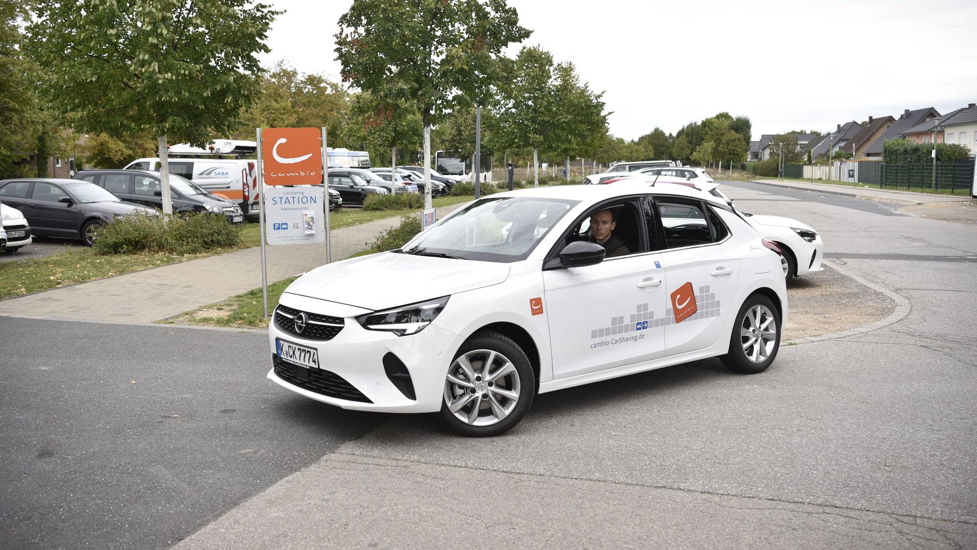 Ein Mann fährt mit einem weißen Auto auf einem Parkplatz. Im Hintergrund sind Bäume und geparkte Autos zu erkennen.