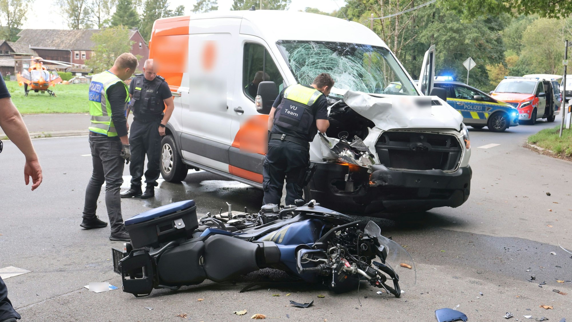 Ein Motorrad liegt nach einem Unfall auf der Straße. Im Hintergrund ist ein Transporter zu sehen, dessen Vorderseite zerbeult und Frontscheibe kaputt ist. Mehrere Polizisten stehen neben den Fahrzeugen. Auf einer Wiese ist ein Rettungshubschrauber gelandet.