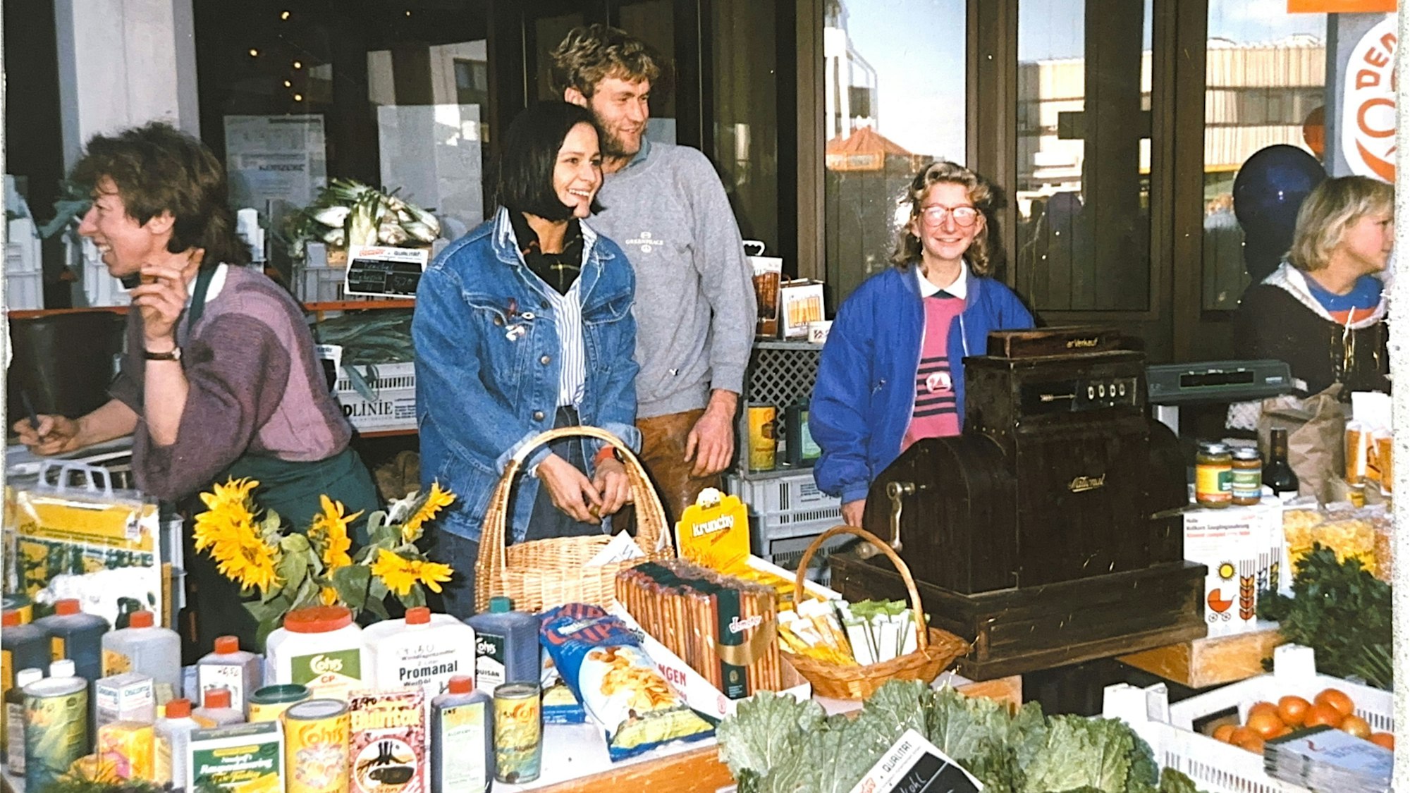 Ein altes Foto, auf dem hinter einem Marktstand mit Dosen, Flaschen und Frischgemüse ein Mann und eine Frau stehen.