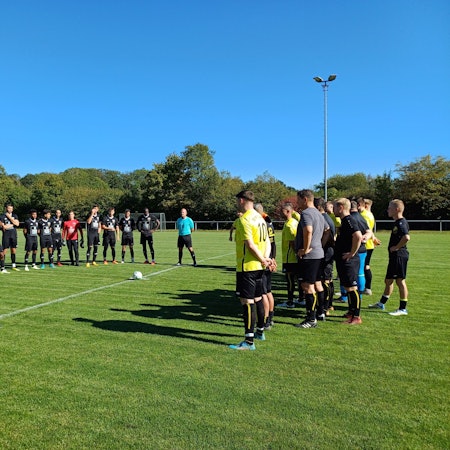 Ein Mann hält eine Ansprache, zwei Fußballteams stehen im Halbkreis um ihn herum und hören zu.